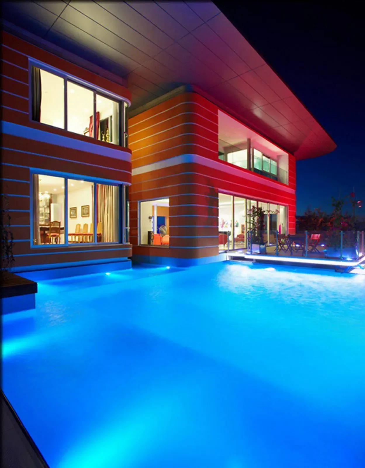 Modern two-story home at night with glowing blue-lit pool and warm interior lights