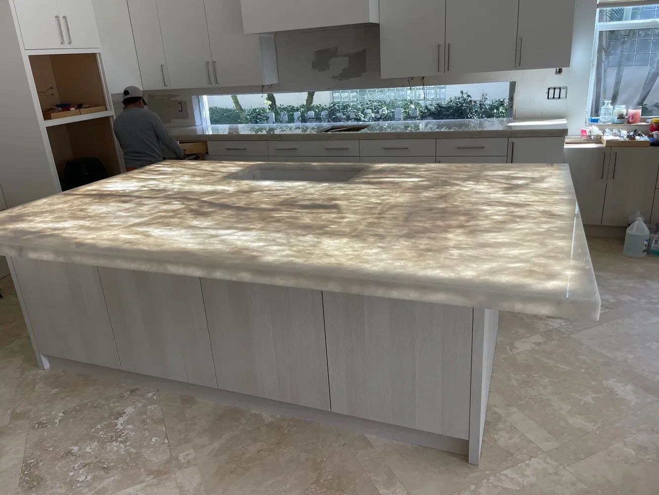 Modern kitchen with a large marble island and light cabinets; a person stands at the left counter.