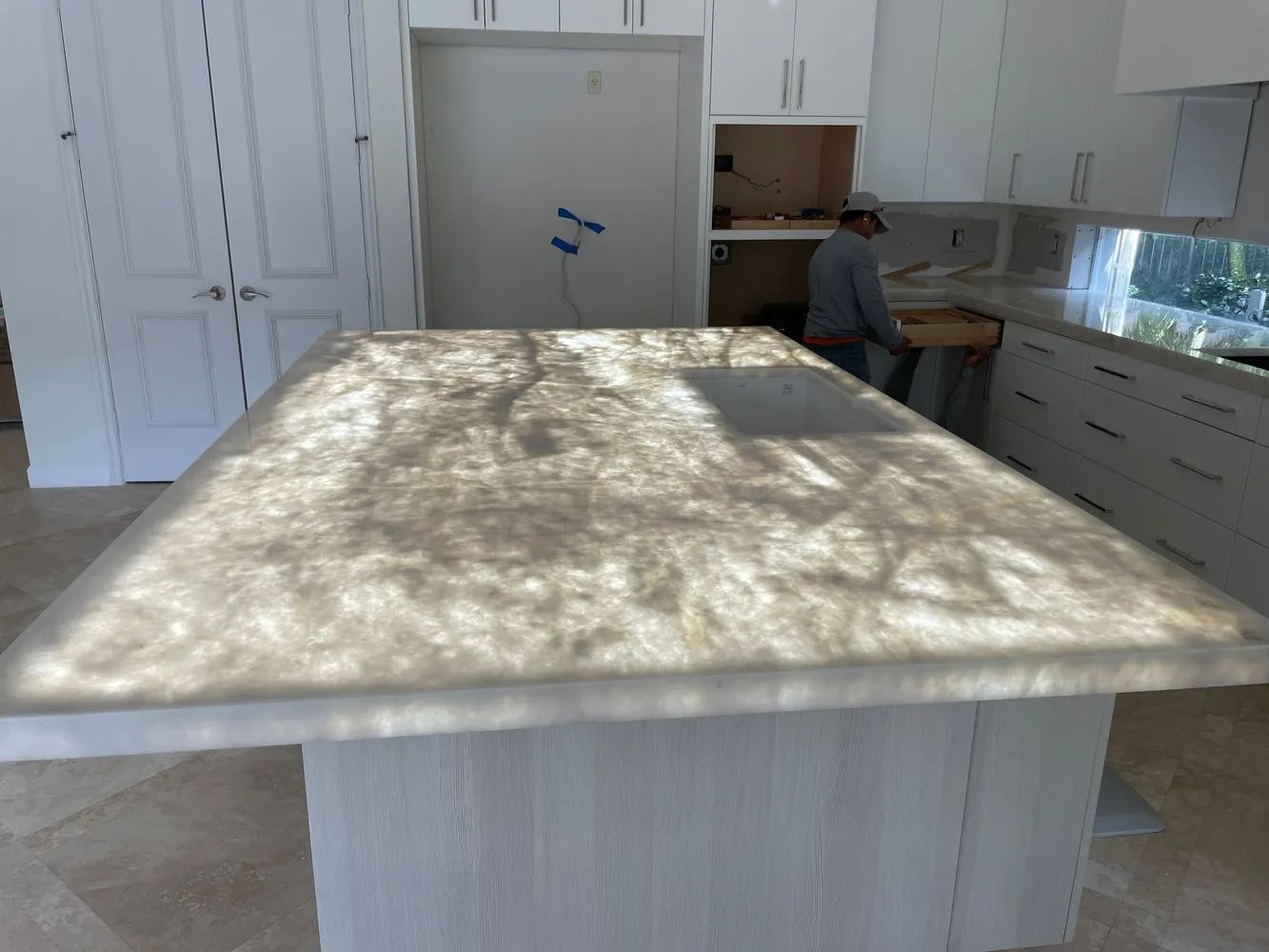 Bright kitchen with white cabinets, large island, and a person working near the back counter.