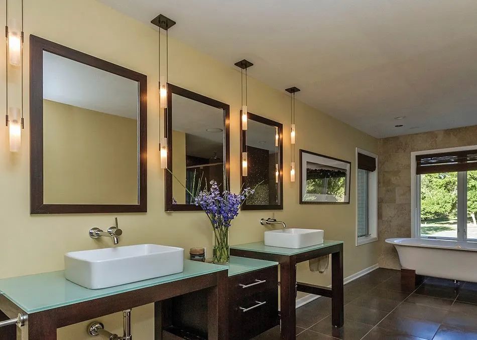 Modern bathroom with two sinks, mirrors, pendant lights, and a bathtub by a window.