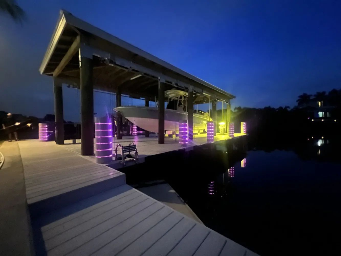 Illuminated lakeside dock with purple and yellow lights at dusk, reflected in calm water