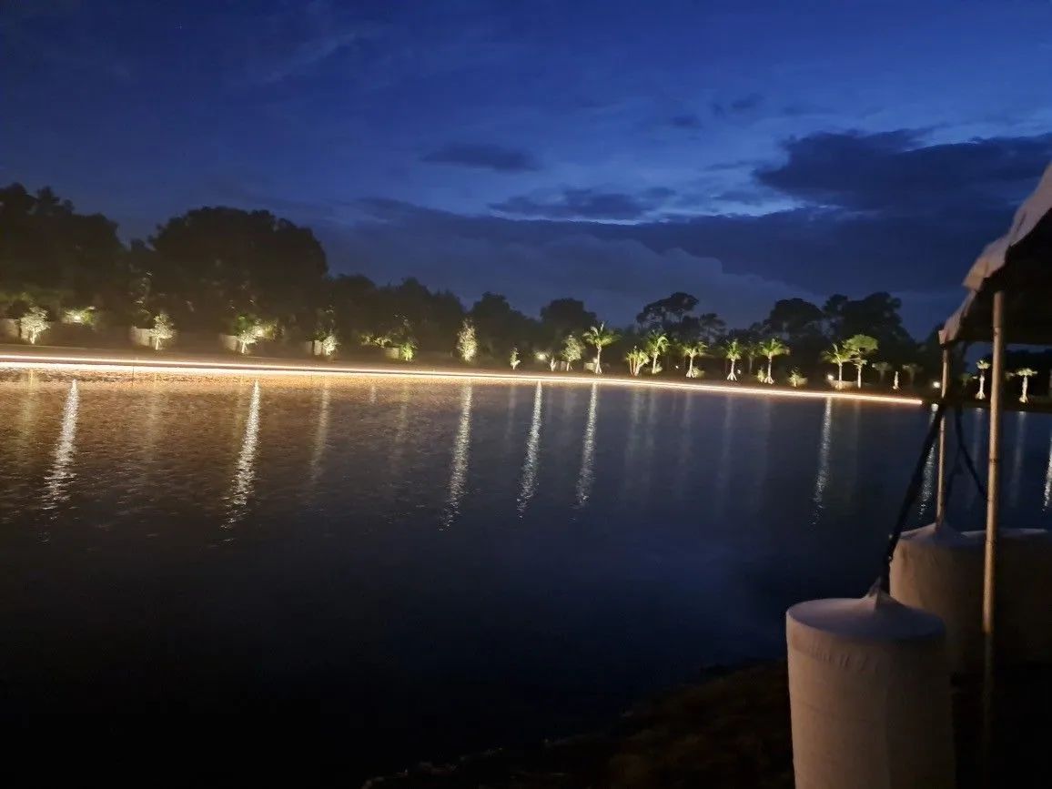 Nighttime lakeside view with glowing shoreline lights reflected on calm water and a boat edge in the foreground