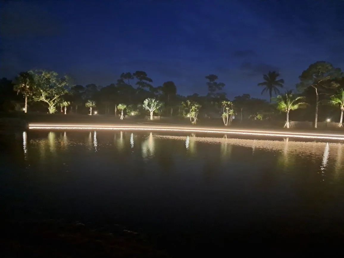 Nighttime lakeside scene with glowing shoreline lights reflected on calm water under a dark blue sky
