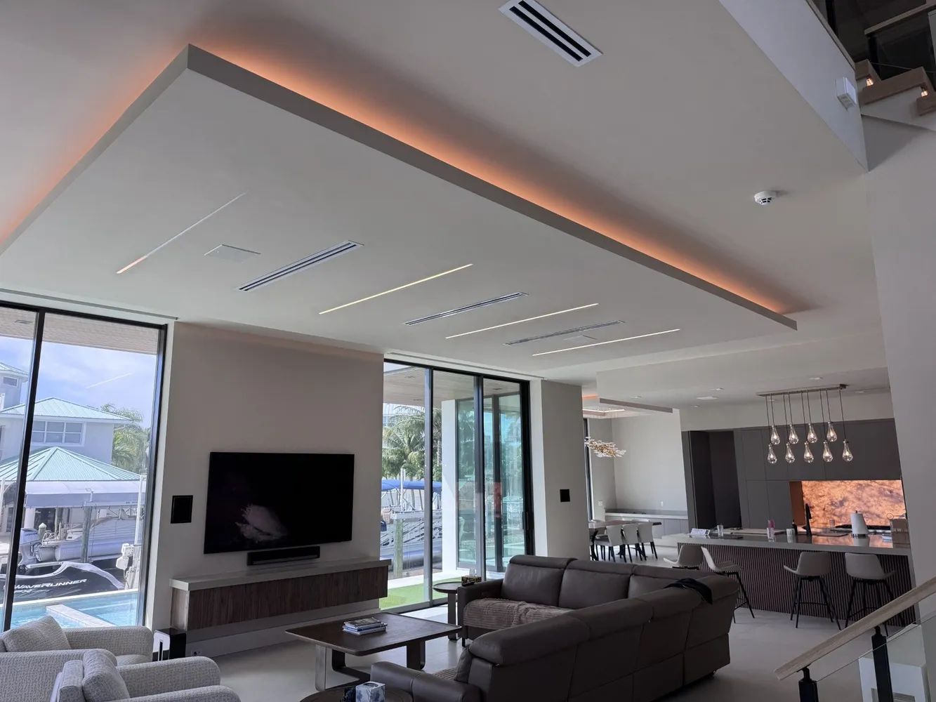 Modern living room with large windows, gray sectional, wall-mounted TV, and open kitchen/dining area