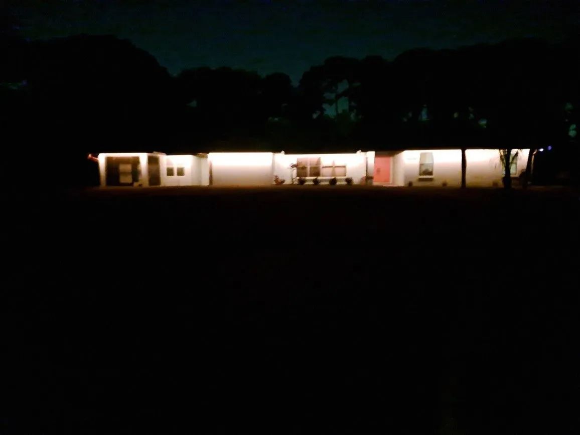 A warmly lit single-story house at night, seen from a dark yard with trees silhouetted above.