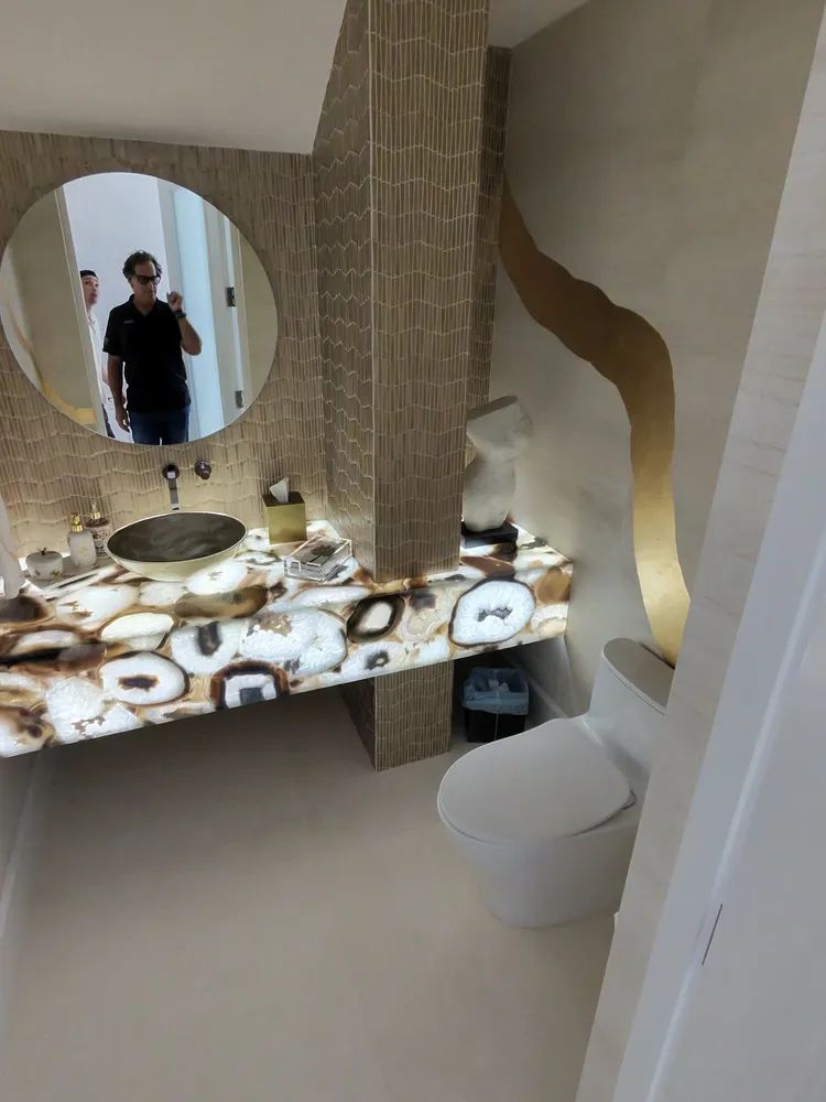 Modern bathroom with patterned stone sink, round mirror, white toilet, and a person reflected in the mirror