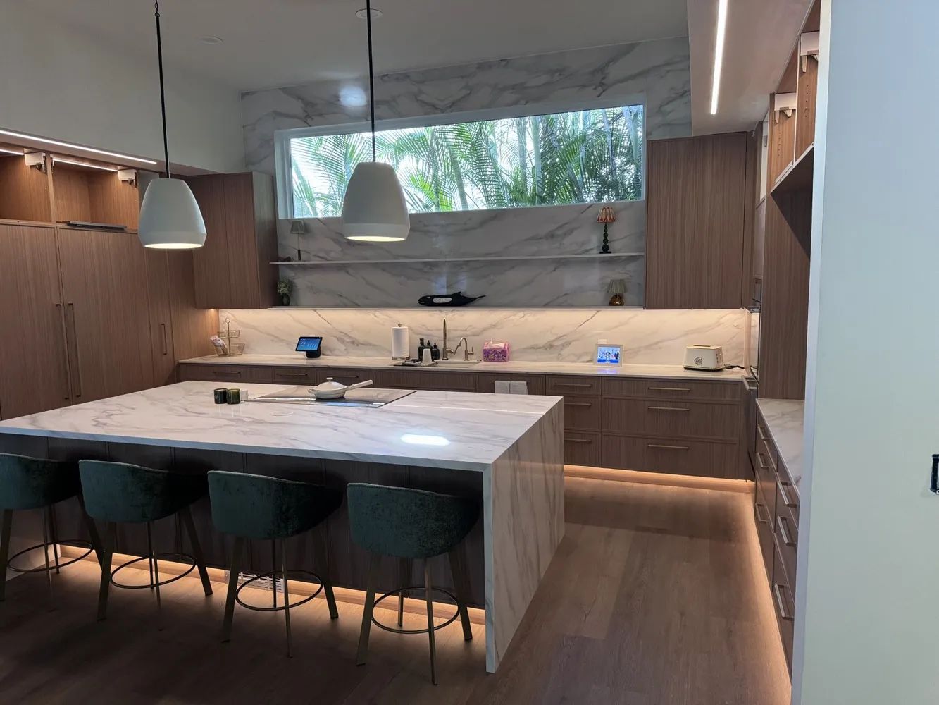 Modern kitchen with a large marble island, black stools, wood cabinets, and a wide window above the sink.