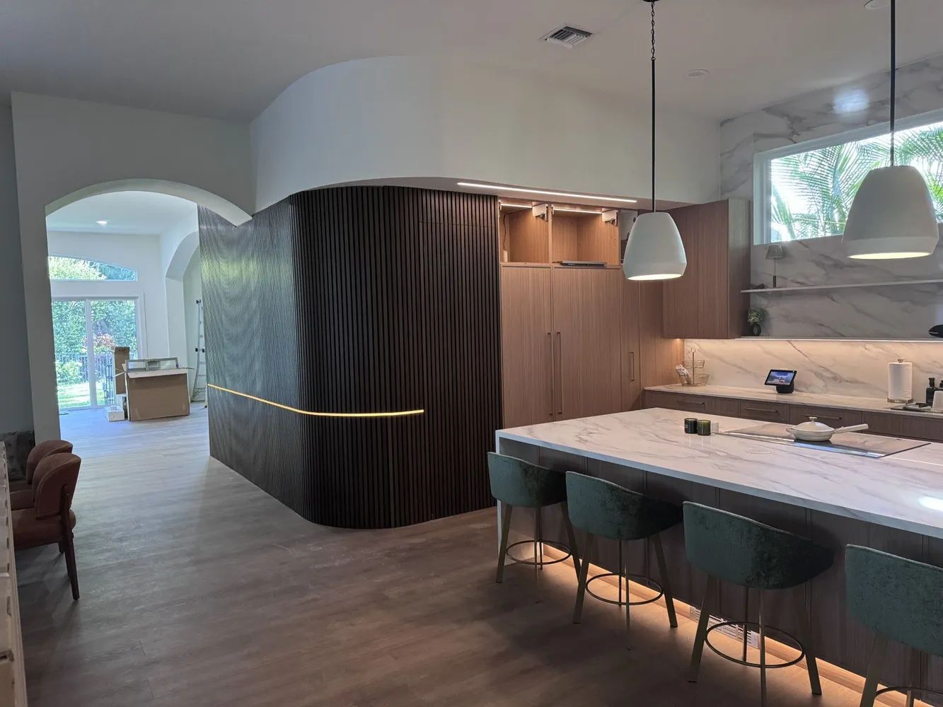 Modern kitchen with marble island, teal stools, wood cabinetry, and curved dark partition in open-plan space