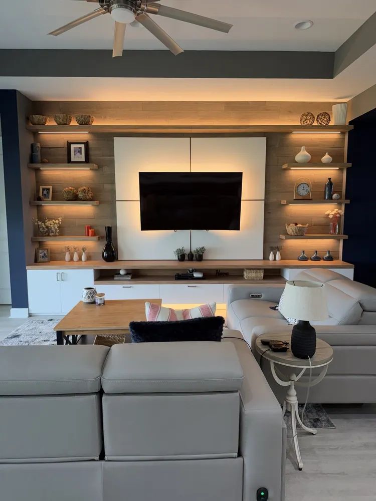 Modern living room with wall-mounted TV, built-in shelves, ceiling fan, and a gray sectional sofa.