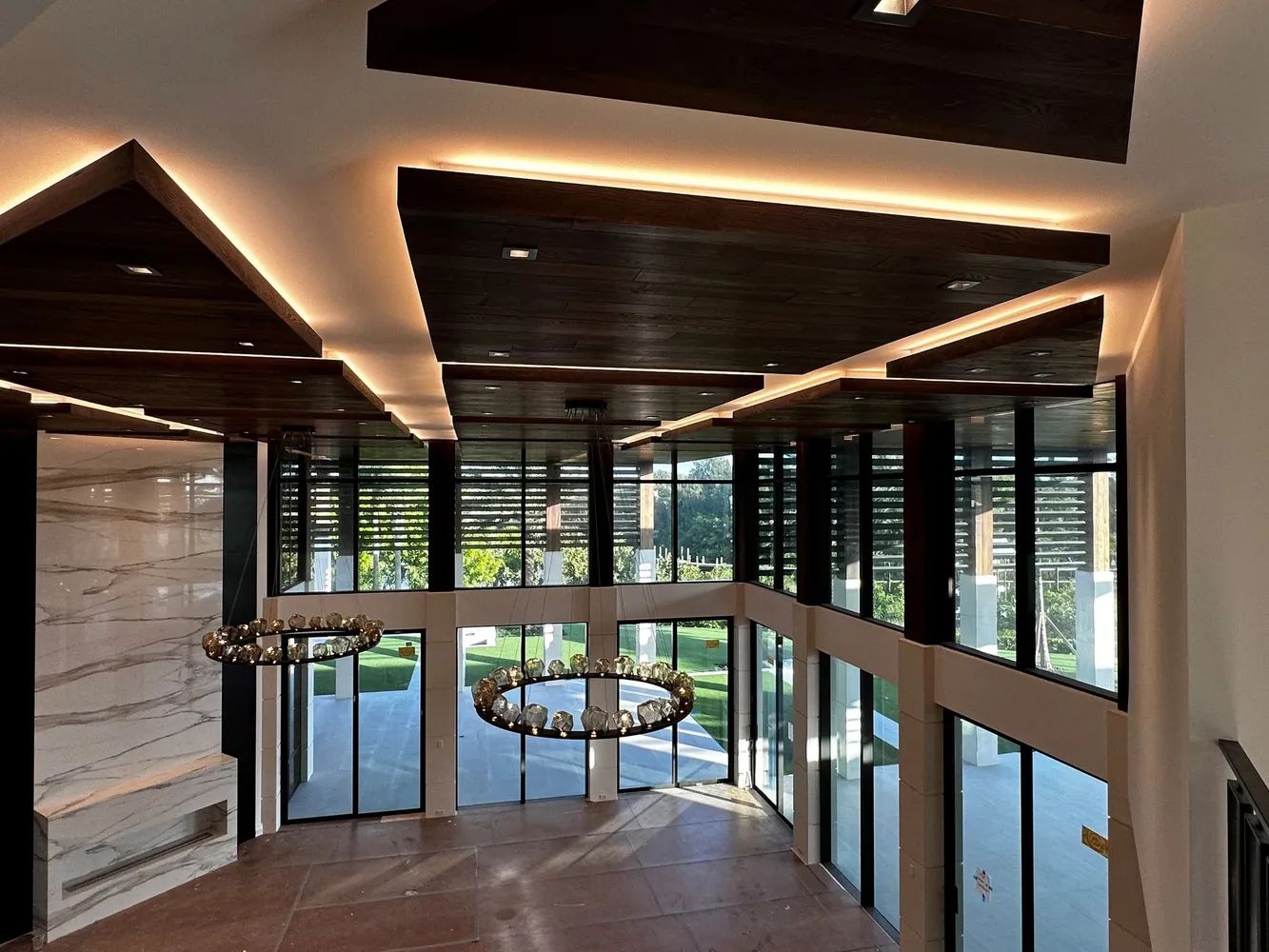 Modern interior with curved staircase, hanging chandeliers, and large windows overlooking greenery