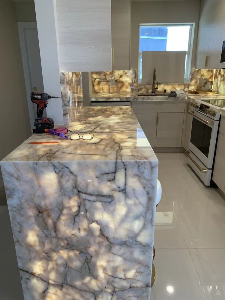 Modern kitchen with glossy white cabinets, marble island, and stone backsplash lit by under-cabinet lights