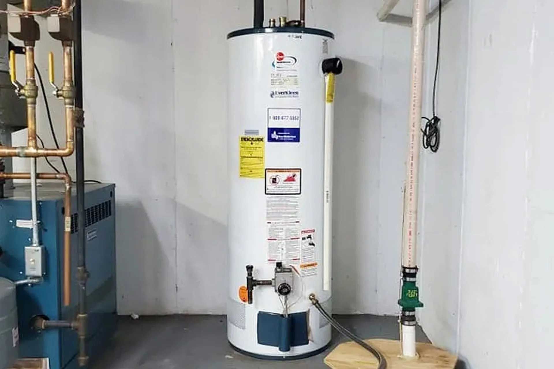 A tall, white water heater in a basement setting with plumbing and pipes.