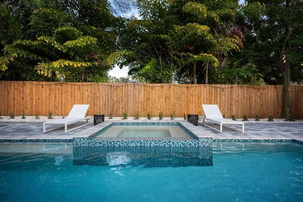 A large swimming pool with two lounge chairs next to it and a wooden fence.
