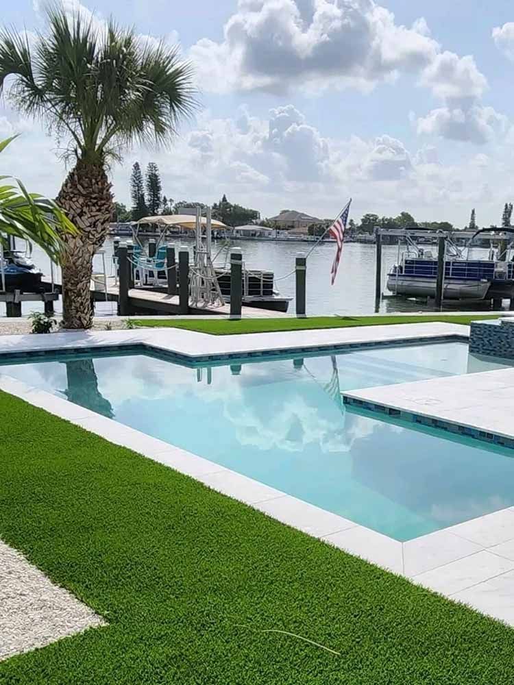 A large swimming pool surrounded by grass and a dock with boats docked in the background.