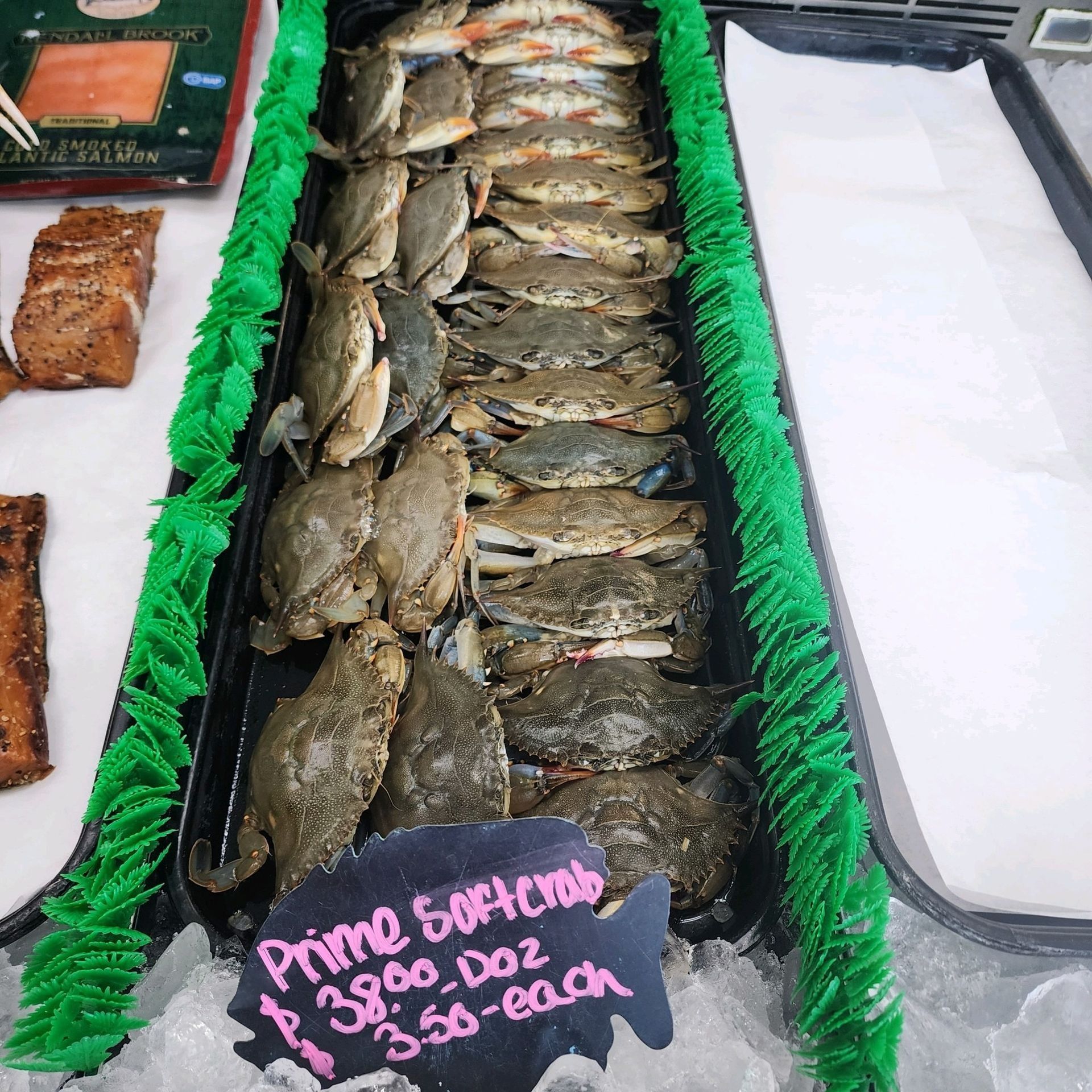 A tray of fresh, raw blue crabs on ice at a seafood market, with a chalkboard sign listing prices per crab and dozen.