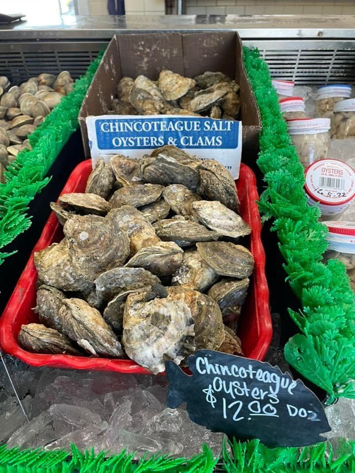 A red basket and cardboard box of Chincoteague salt oysters and clams on ice, with a sign pricing them at $12.00 per dozen.