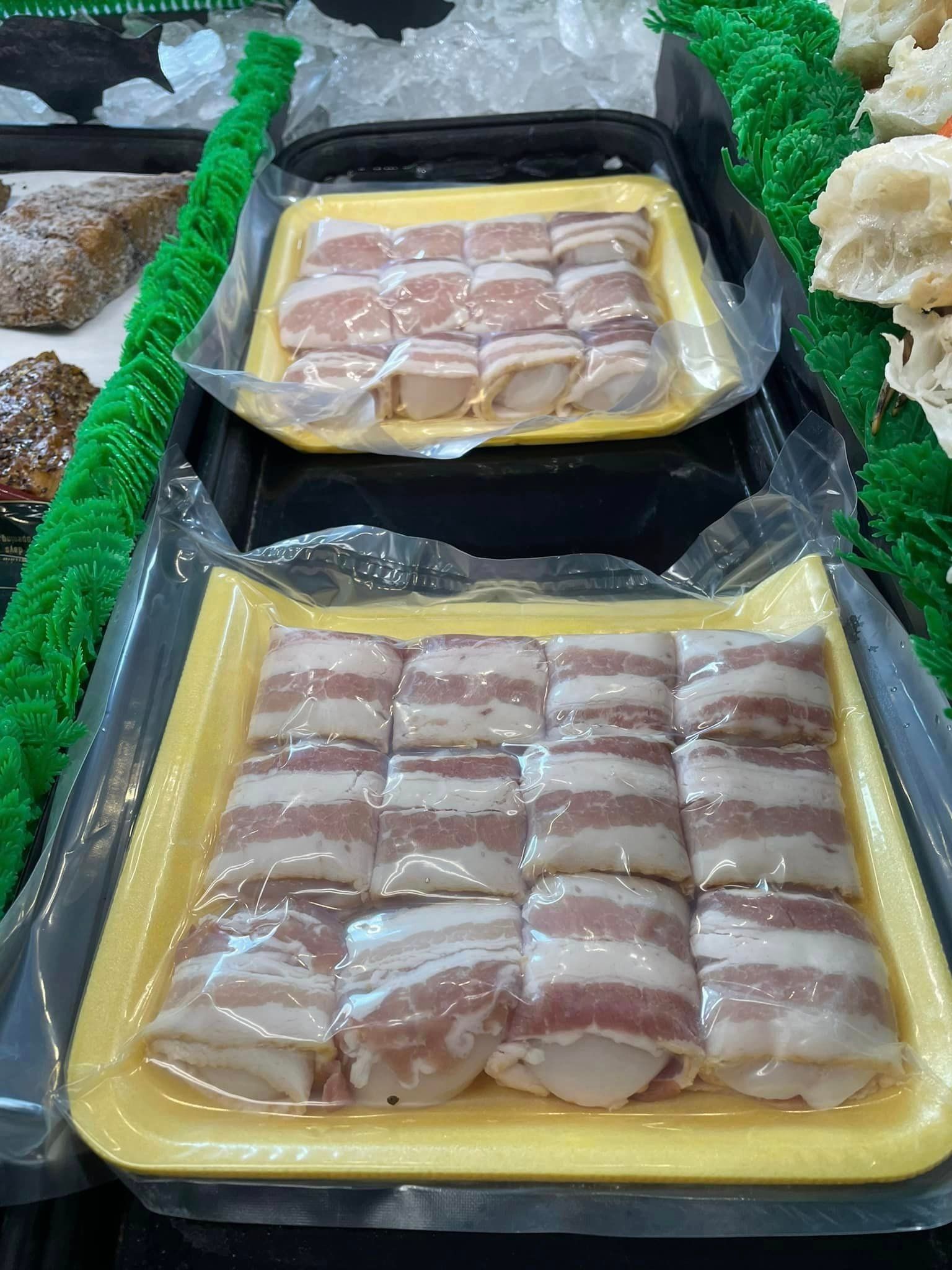 Two plastic-wrapped yellow trays of raw bacon-wrapped scallops on display at a grocery store meat counter.