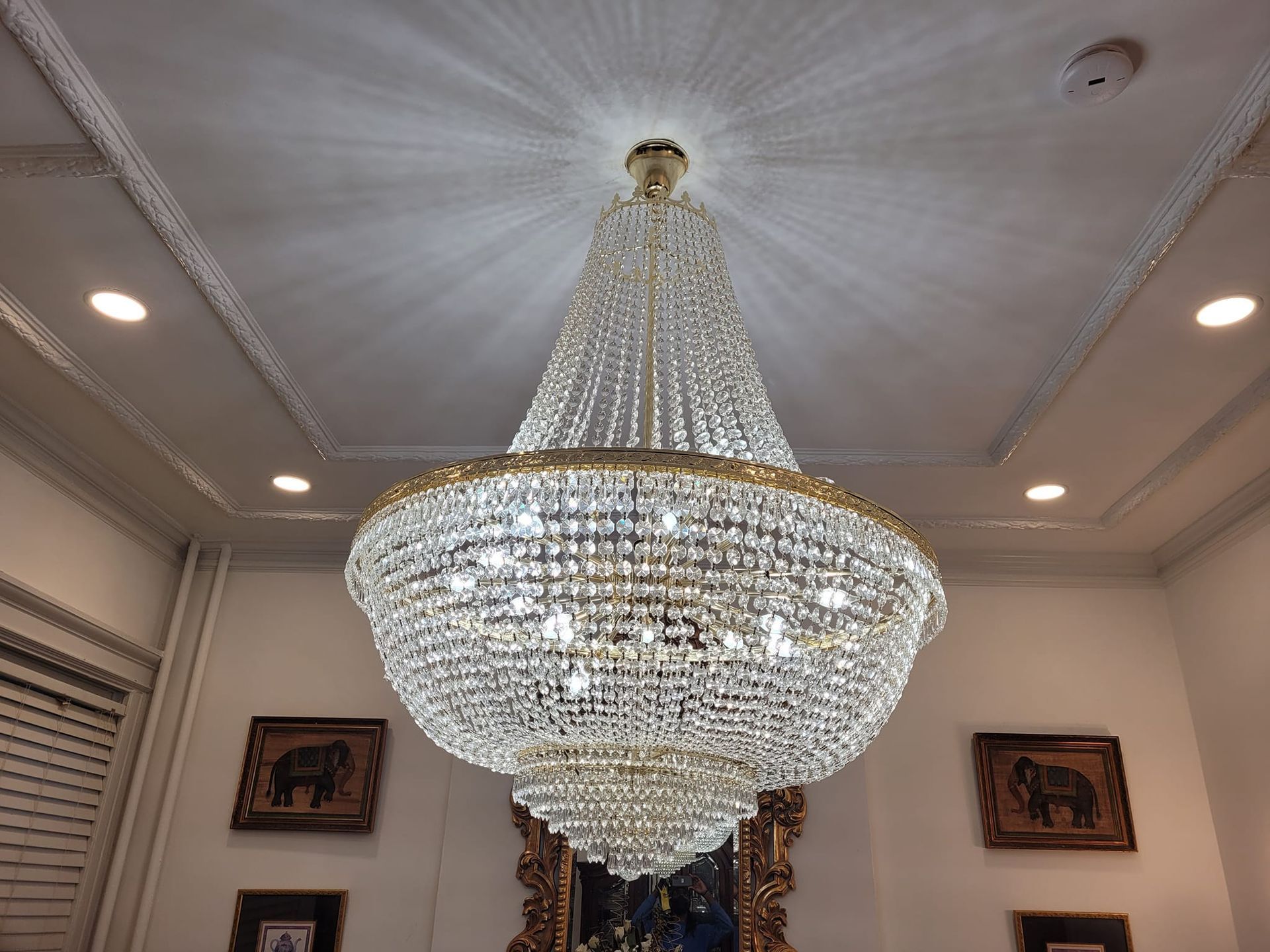 A large crystal chandelier hangs from a decorative ceiling, with recessed lighting and framed artwork below.