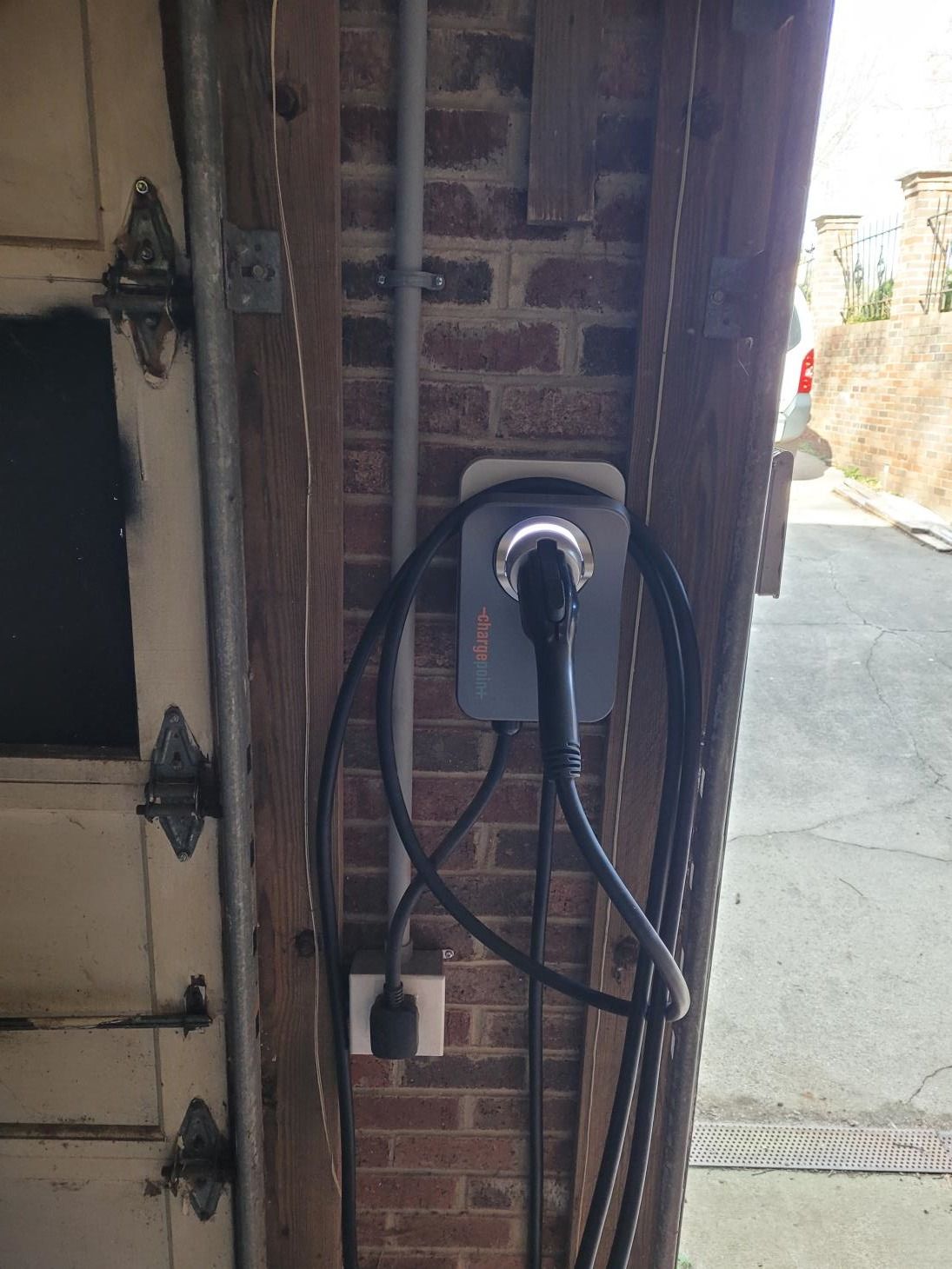 EV charger mounted on a brick wall, near a garage door. A black cable is plugged into the charger.