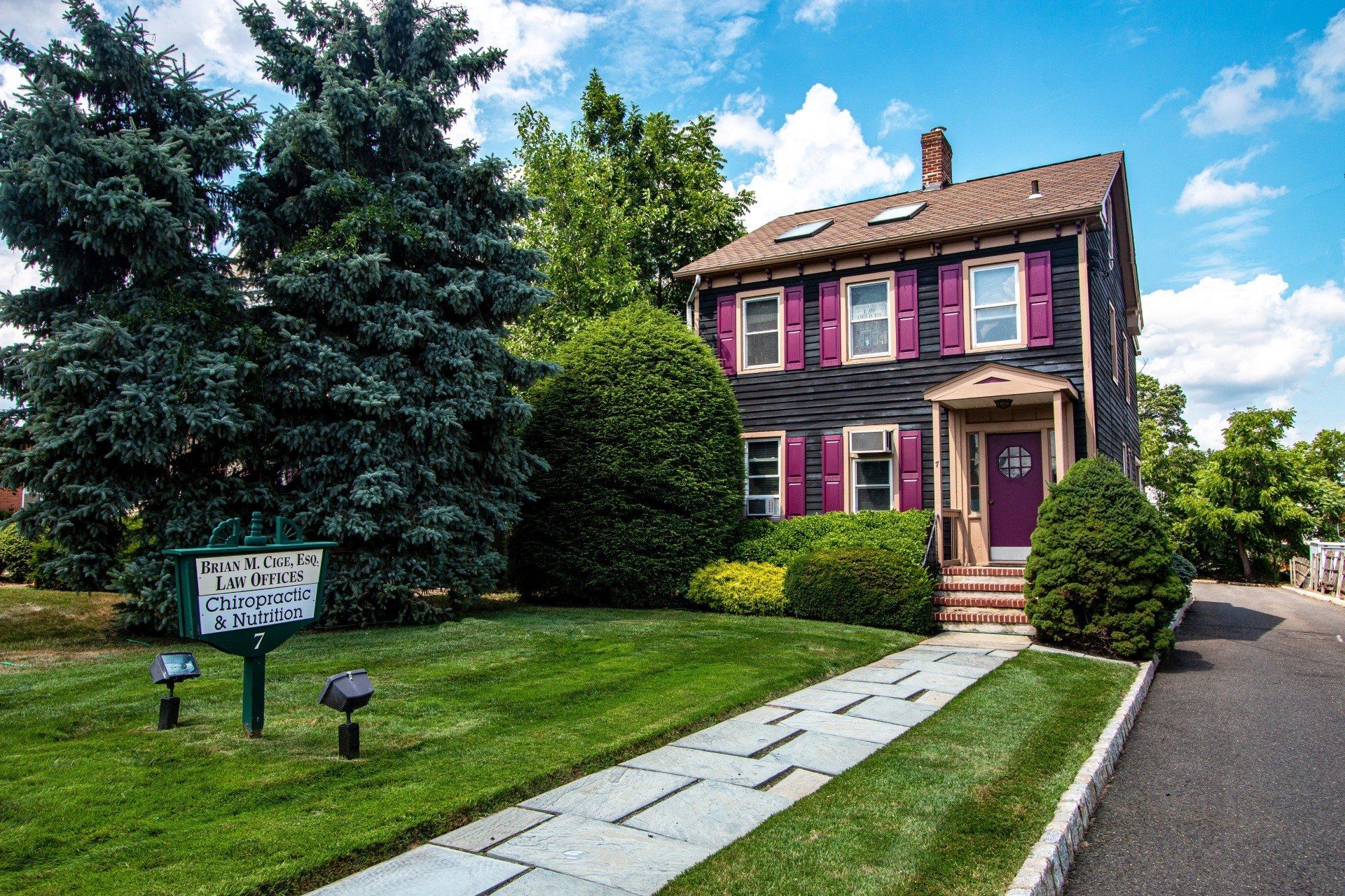 Small pink-and-purple house with a stone walkway, lawn, and large trees beside a road