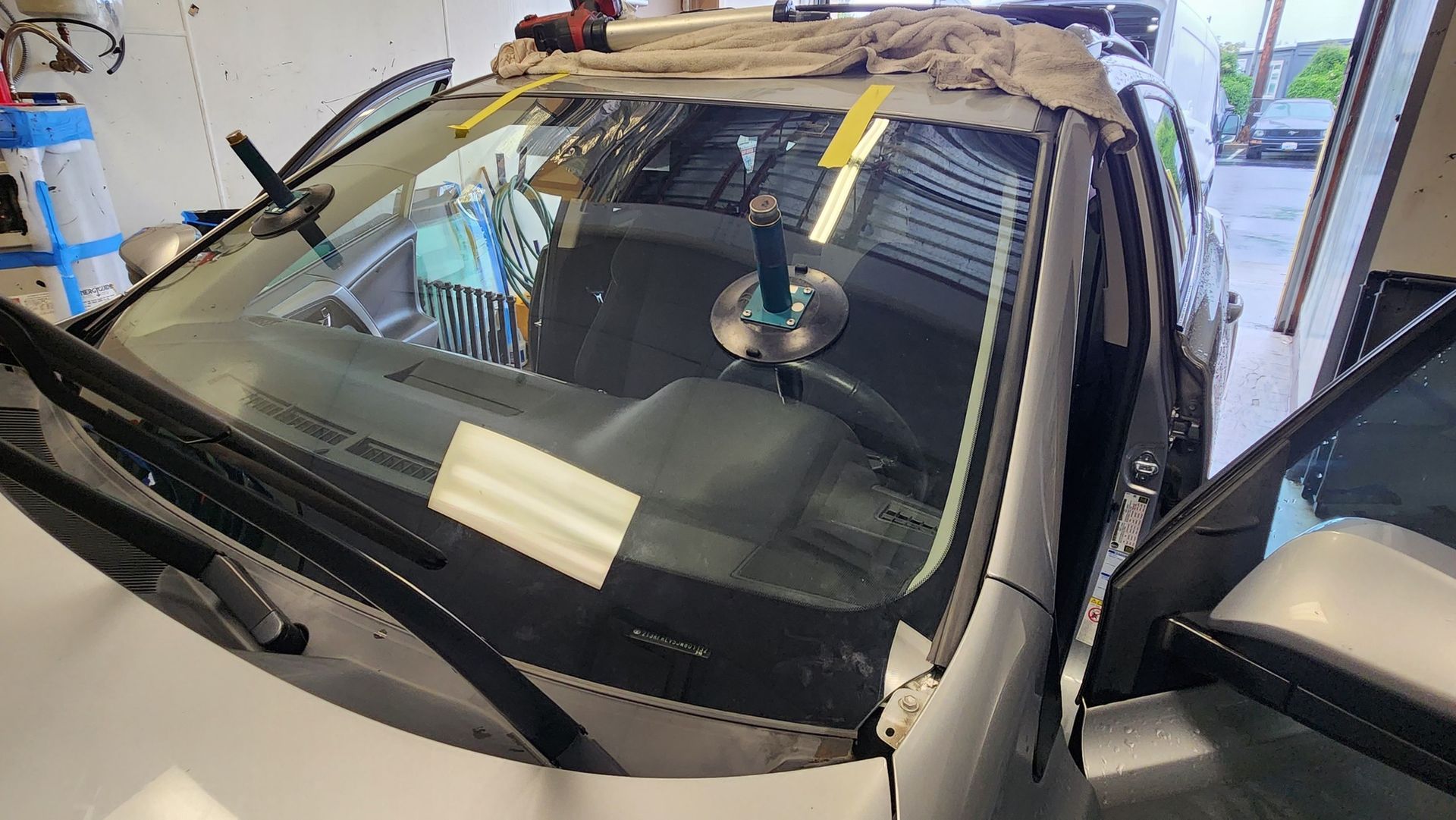 A car windshield being replaced inside a garage with tools, suction cups, and a cloth covering the roof.