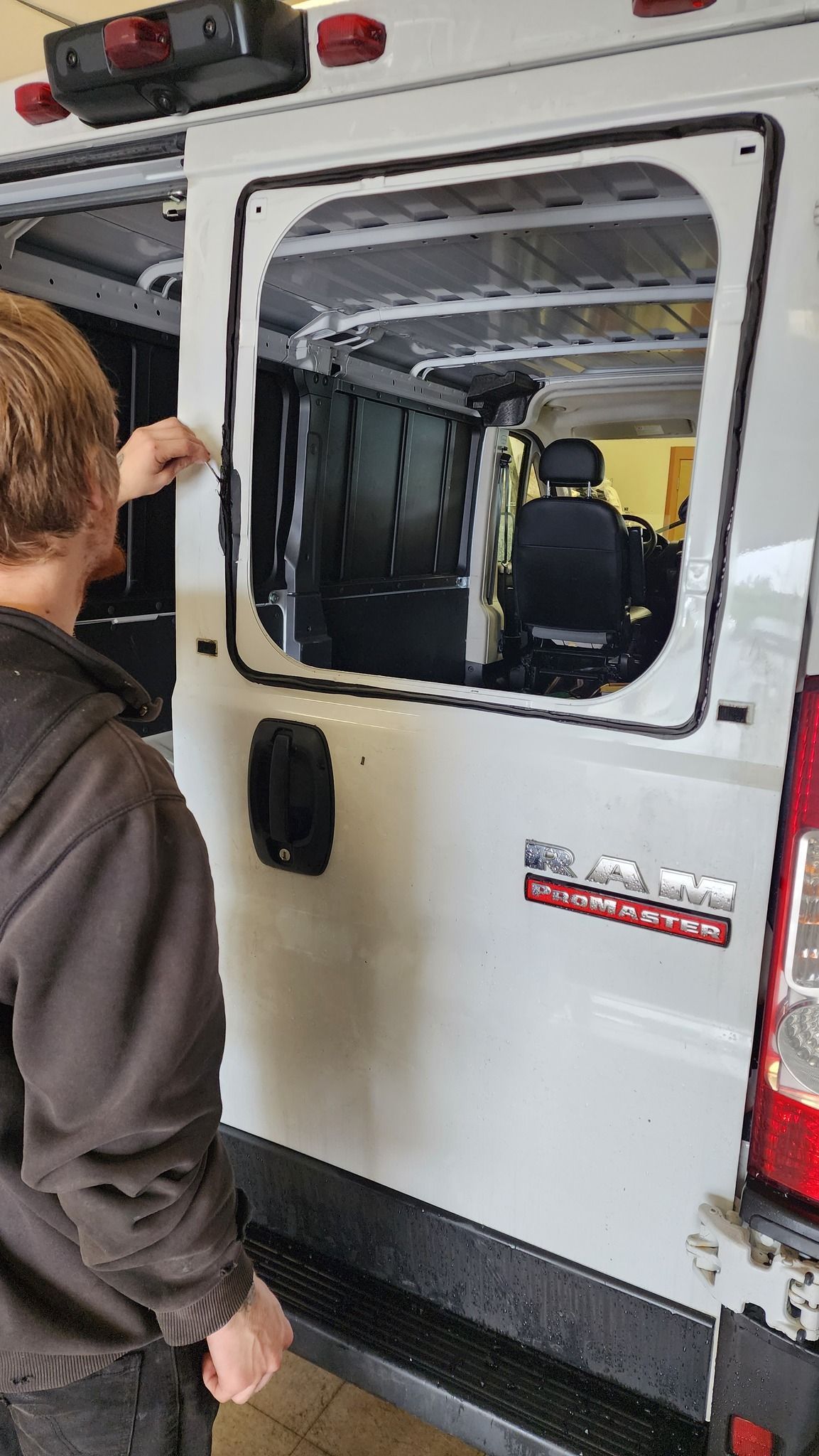 Man installing a window on a white RAM van. Black trim, open cargo area visible inside.