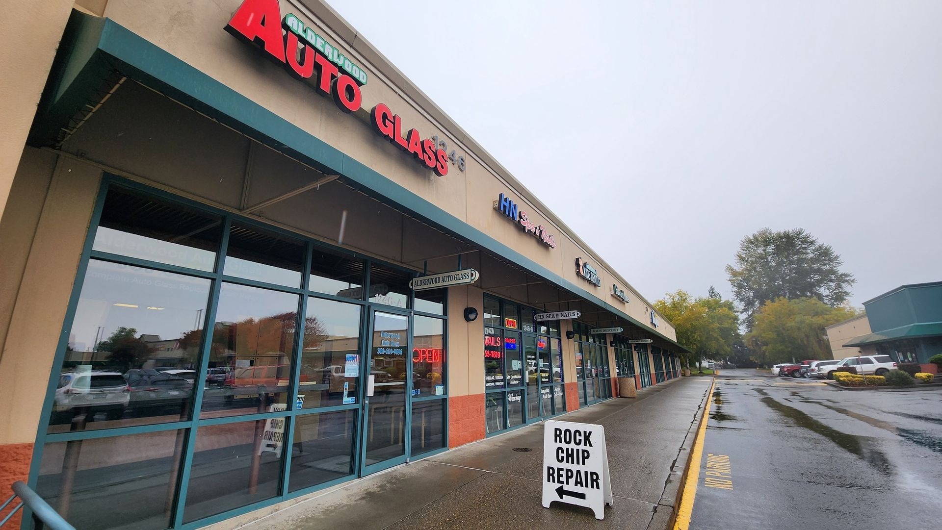 Auto Glass shop and other storefronts in a strip mall, overcast day. A sidewalk and wet street in the foreground.