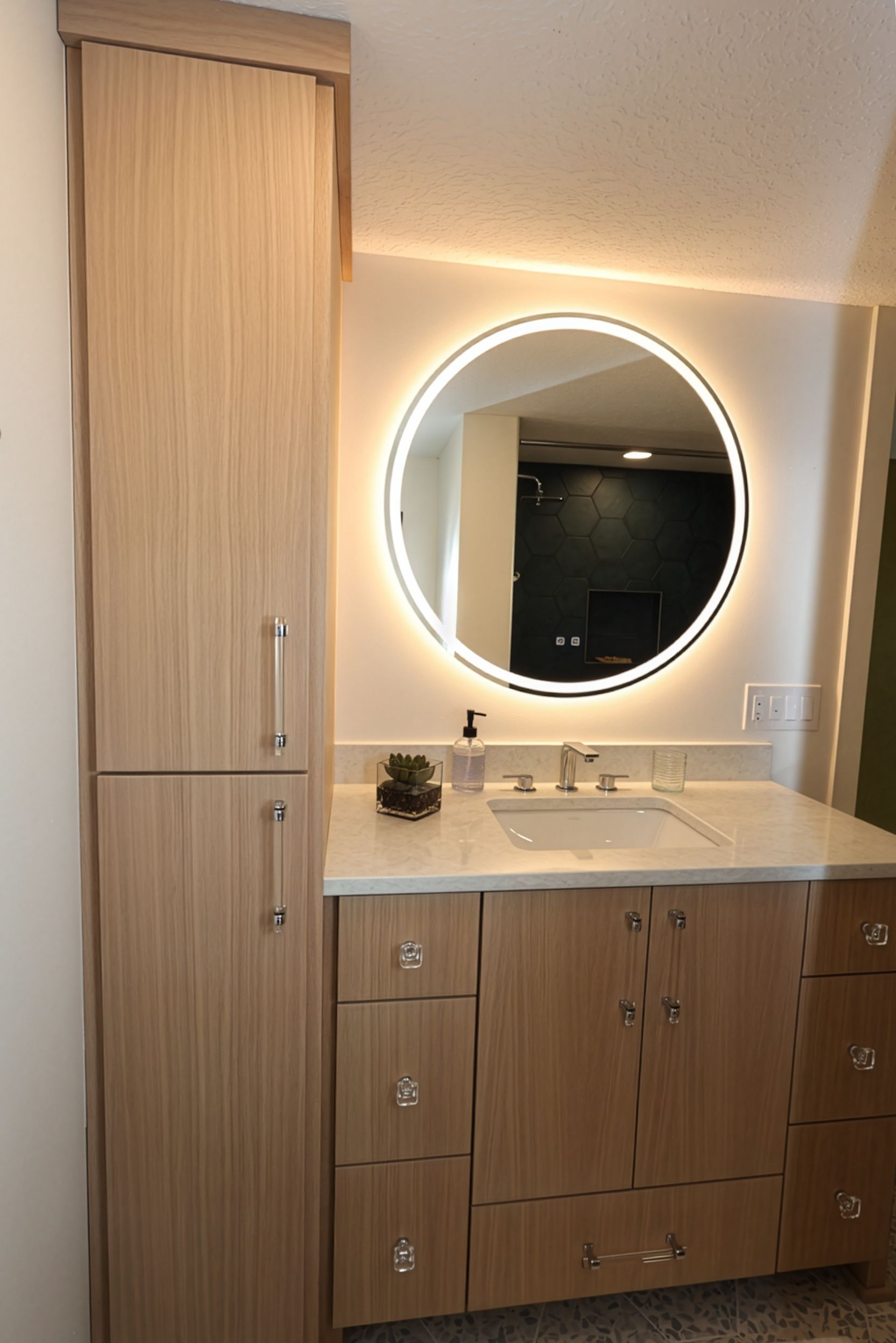 Bathroom vanity with round lit mirror, wood cabinets, and a tall storage cabinet