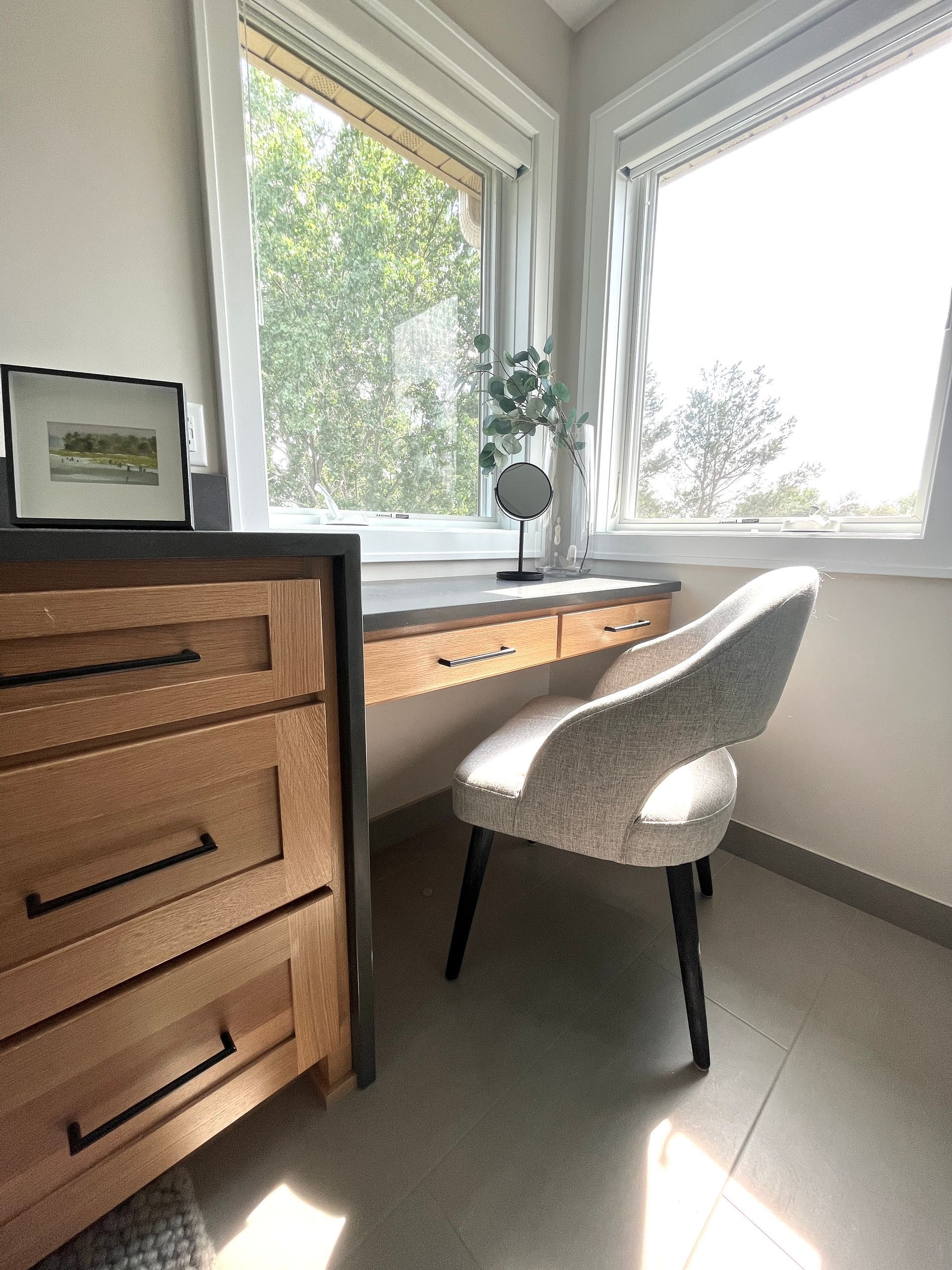 A sunlit corner featuring a wooden desk with a textured light-colored chair, placed between two large windows.