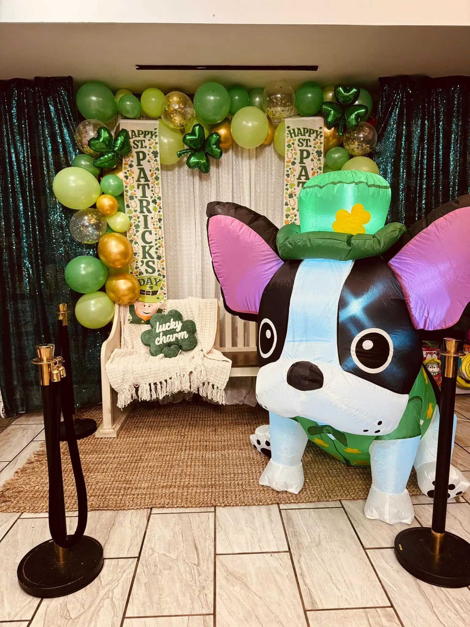 A St. Patrick's Day photo backdrop with green balloons, a white bench, and a large inflatable dog wearing a leprechaun hat.