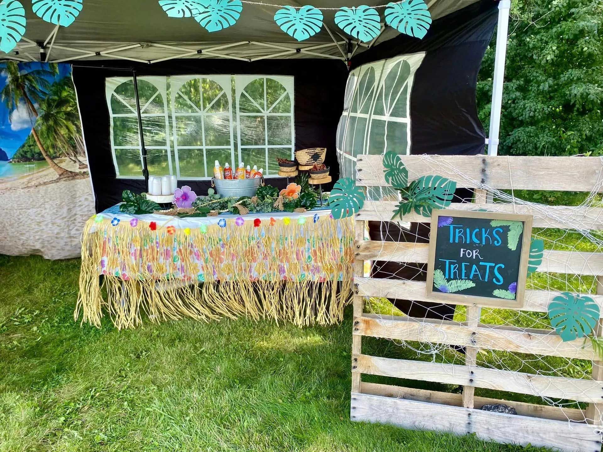 A tropical-themed party booth featuring a straw-skirted table and a wooden pallet sign reading 