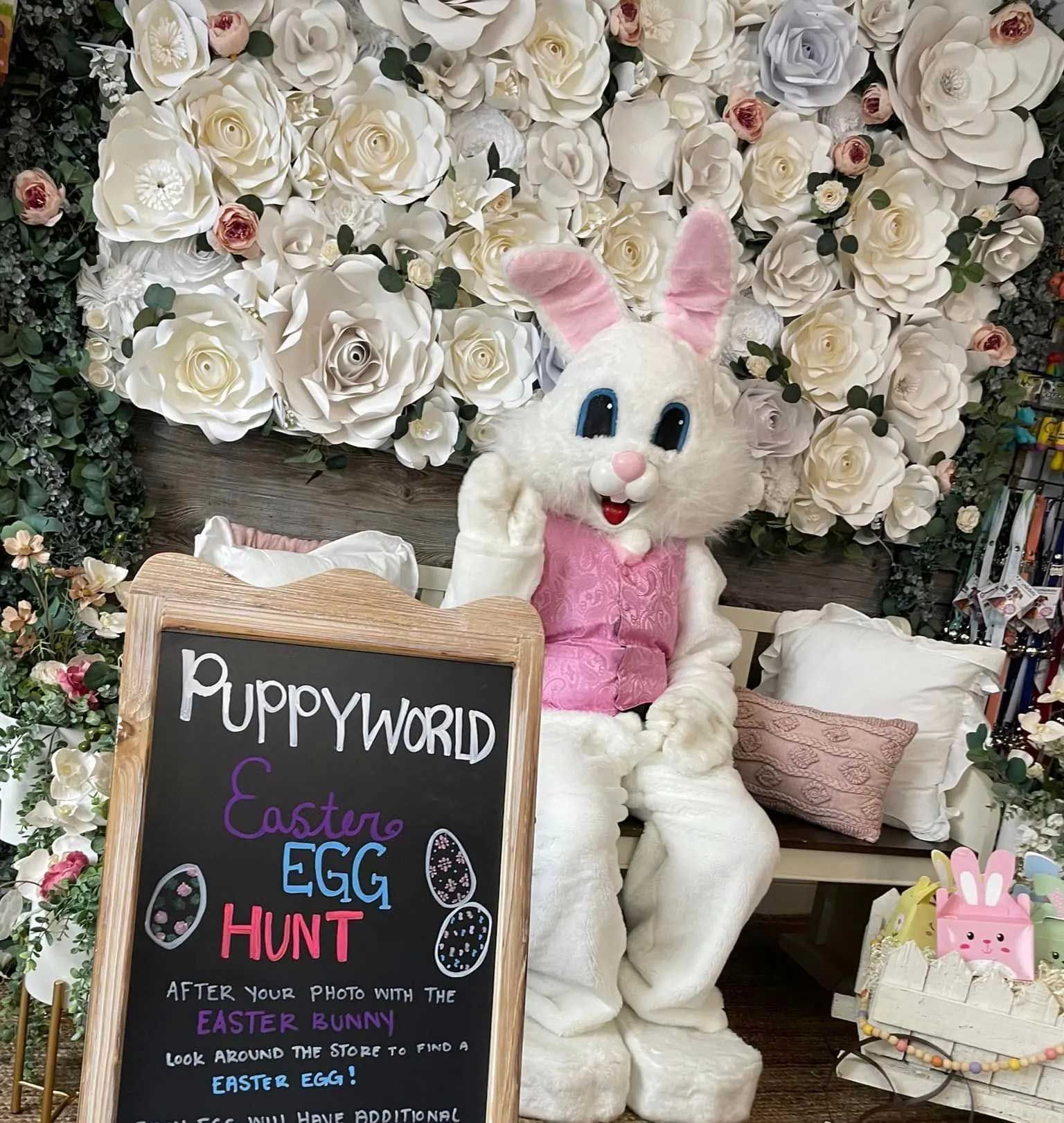 The Easter Bunny waves in front of a flower wall behind a chalkboard sign for a Puppyworld Easter egg hunt.