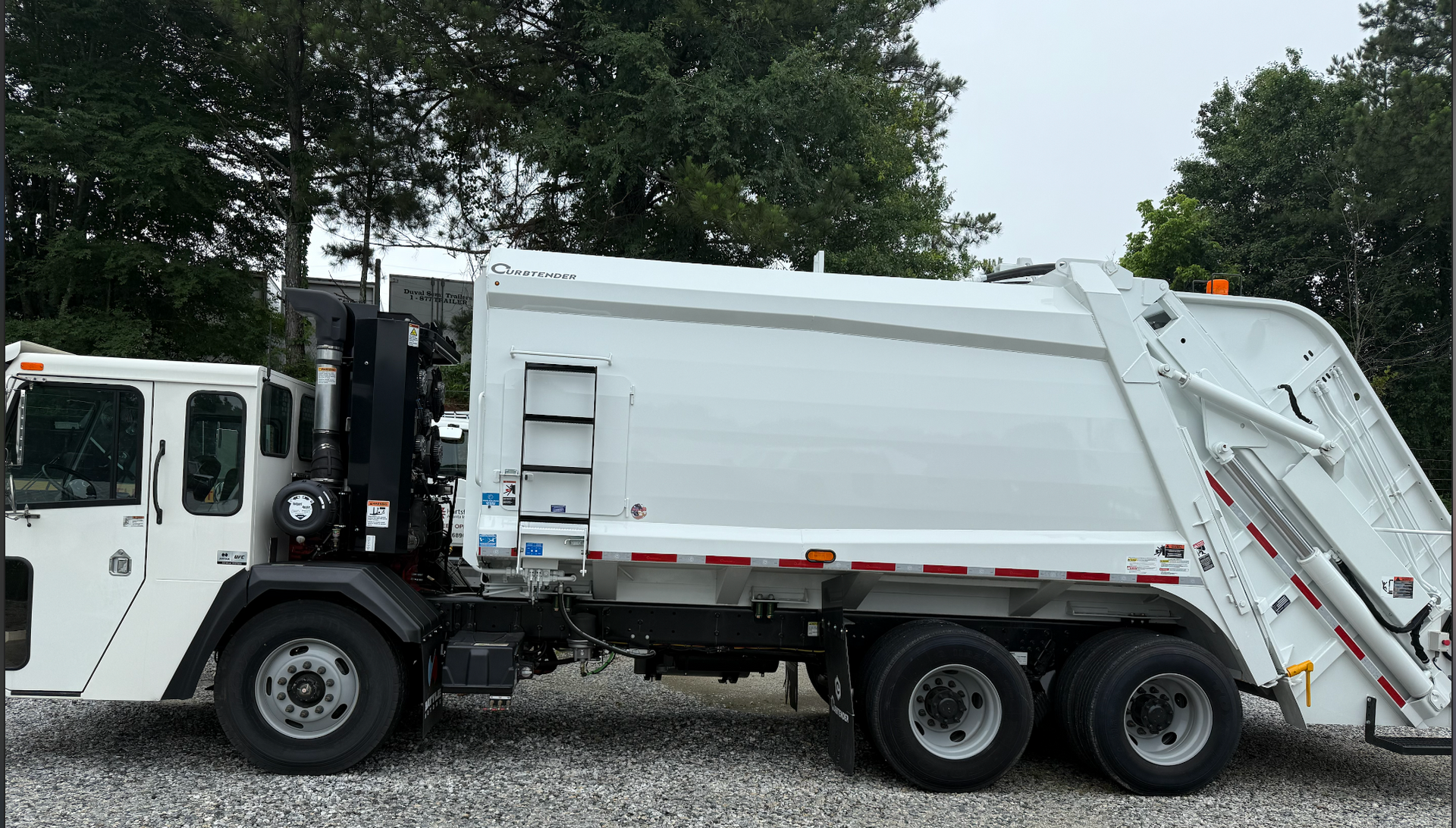 White garbage truck parked on gravel road, trees in the background.