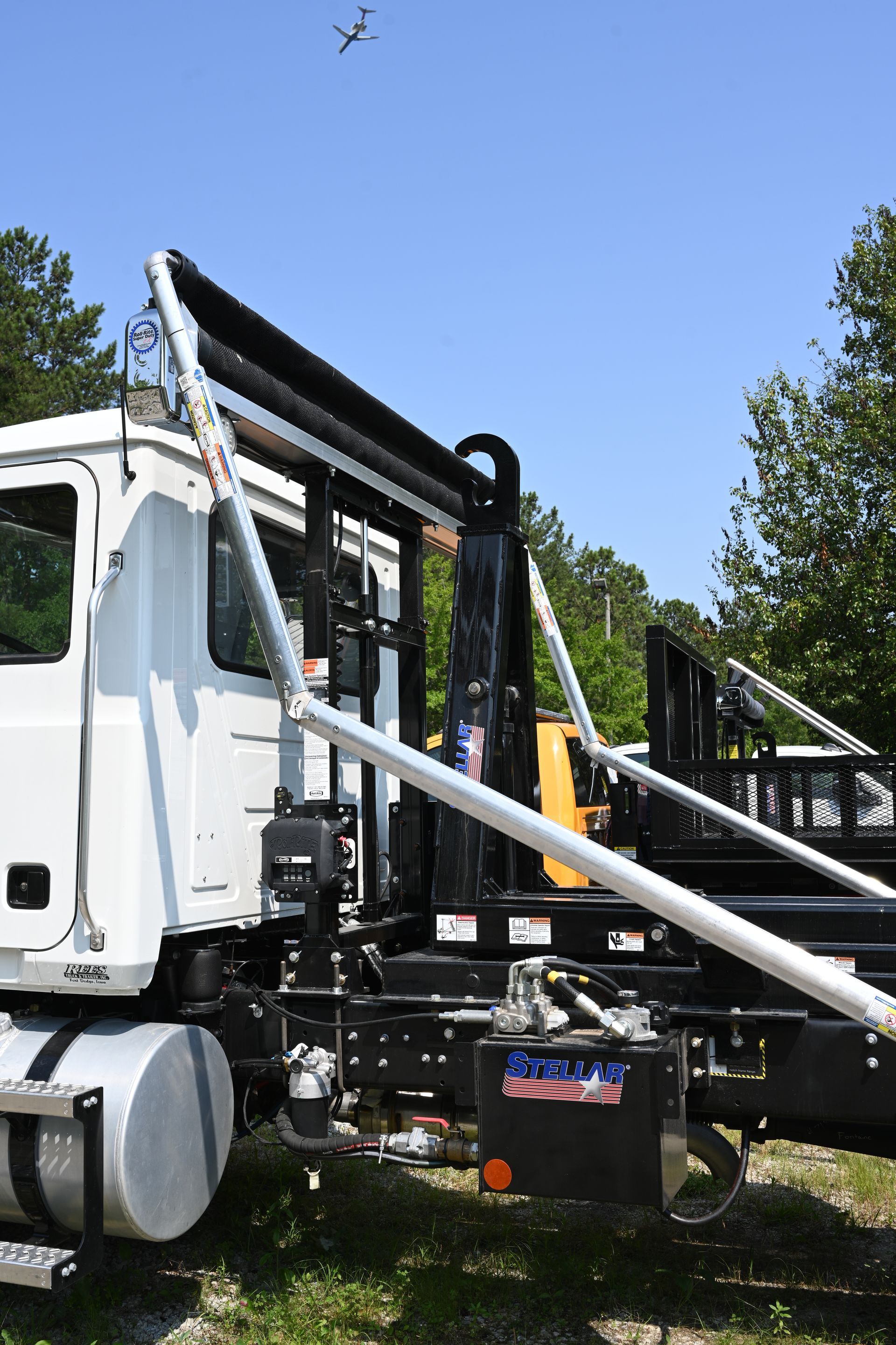 A white dump truck is parked in the grass with a plane flying in the background.