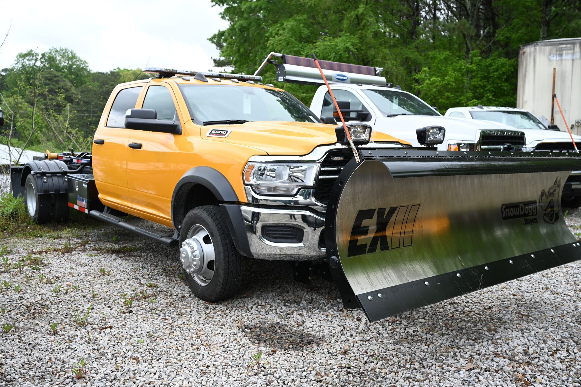 A yellow truck with a snow plow attached to it is parked in a gravel lot.