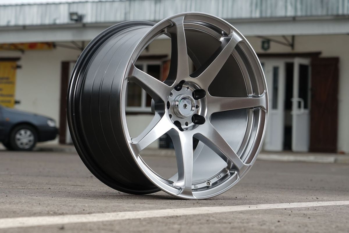 Silver alloy car wheel, seven-spoke design, on asphalt in front of a building.