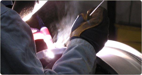 Welder in white protective clothing using a torch to weld on a large metal cylinder, creating bright sparks.