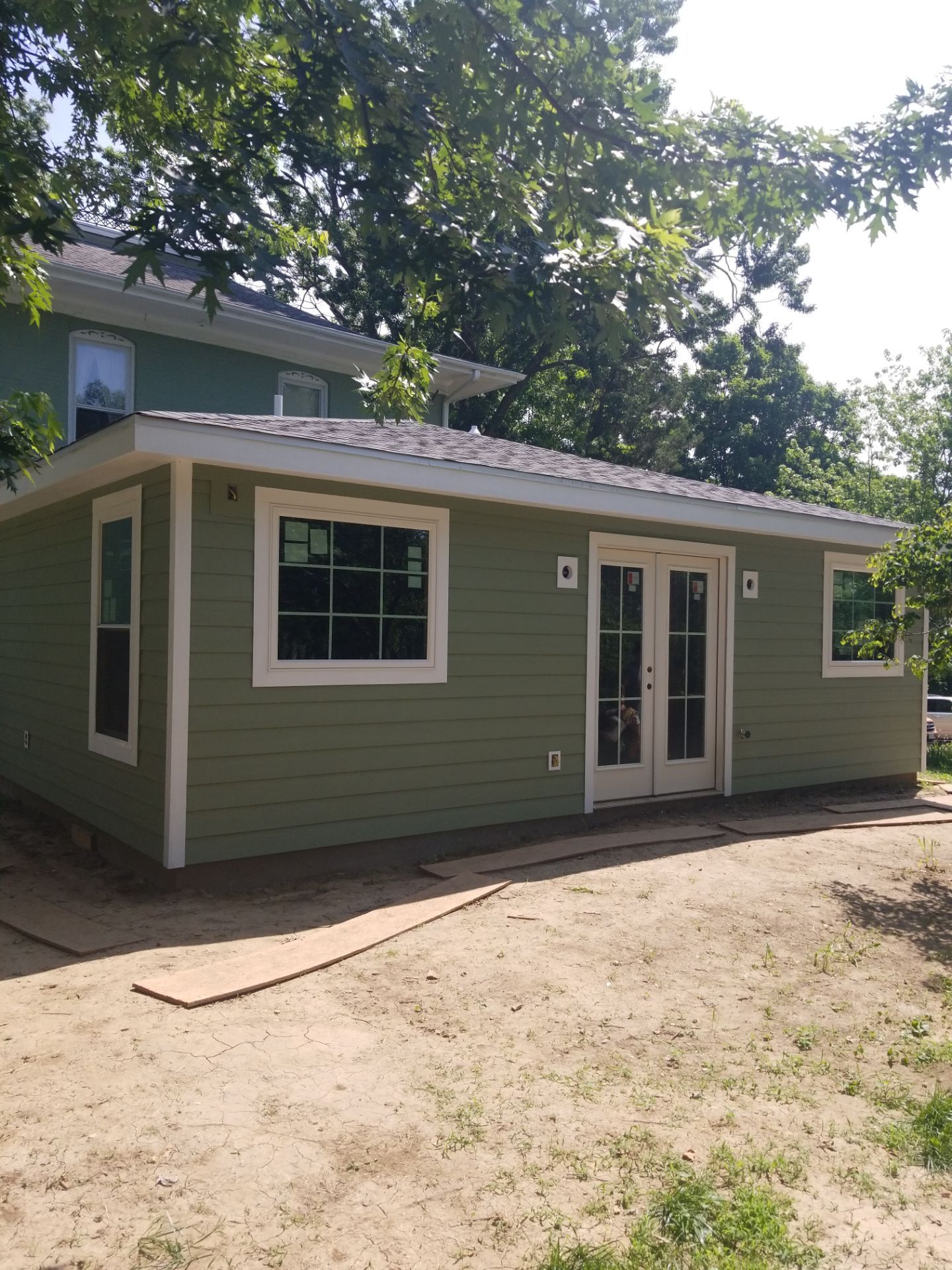 Green, single-story building with white-trimmed windows and double doors; constructed on a dirt lot, tree background.
