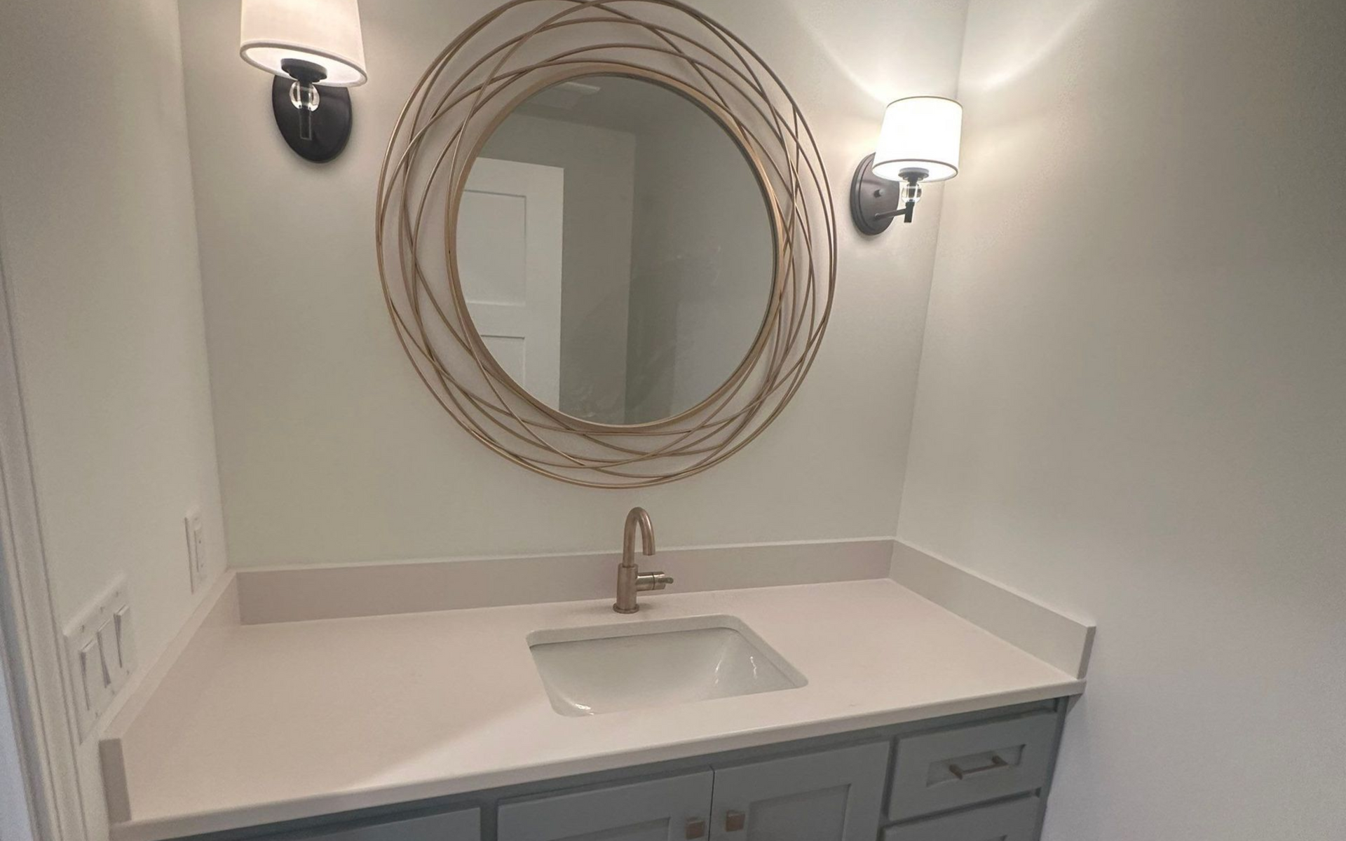 Bathroom with gray cabinets, white countertop, round mirror, and two sconce lights.