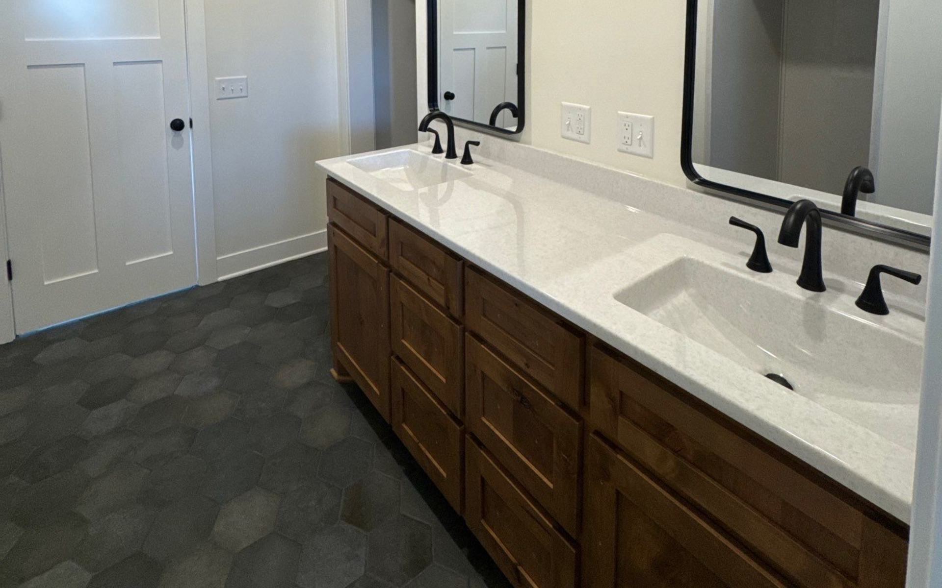 Bathroom with double vanity, white countertop, dark cabinets, black fixtures, and hexagon tile floor.