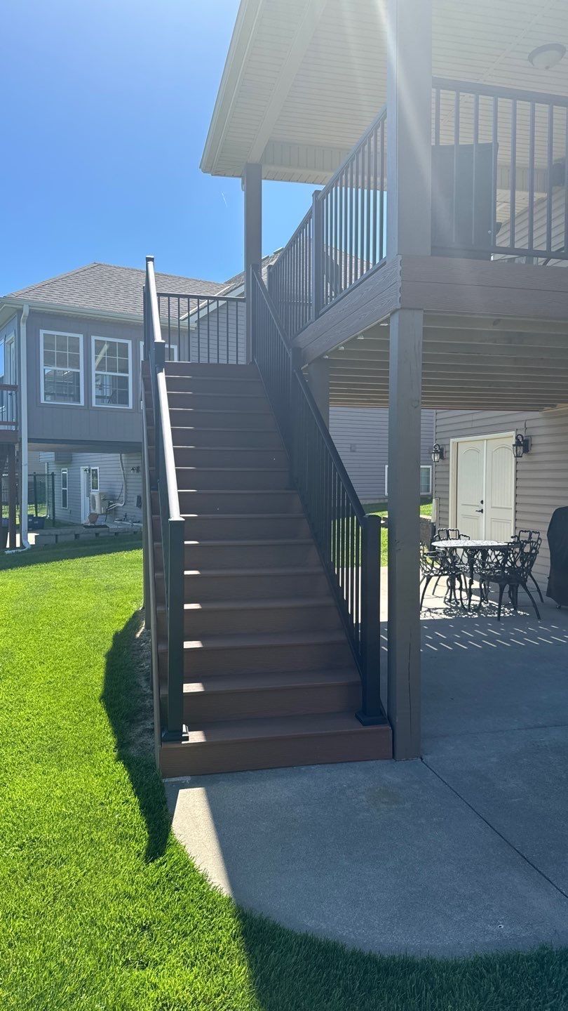 Staircase leading up to a deck with dark railing. Brown stairs, beige siding, green grass, and blue sky.