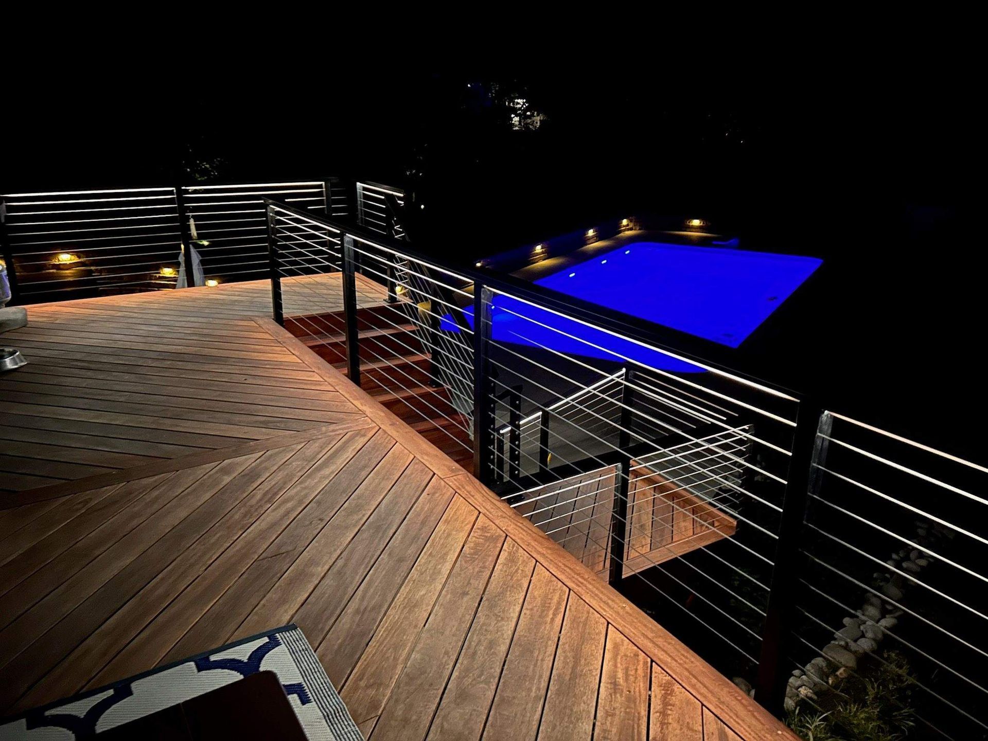 Wooden deck with metal railing overlooking a brightly lit blue pool at night.