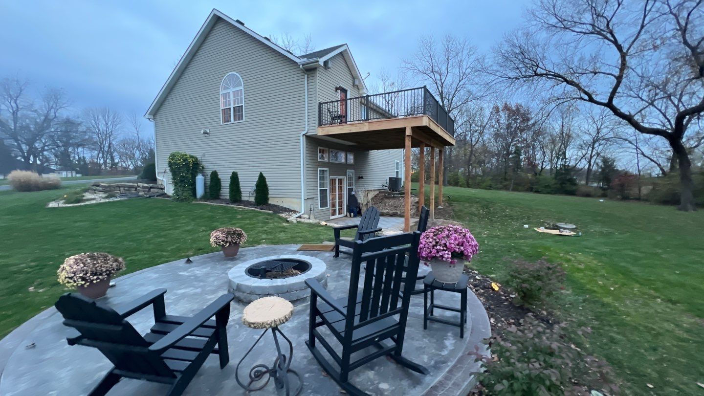 Backyard with a stone patio, fire pit, black chairs, and two-story house with a deck.