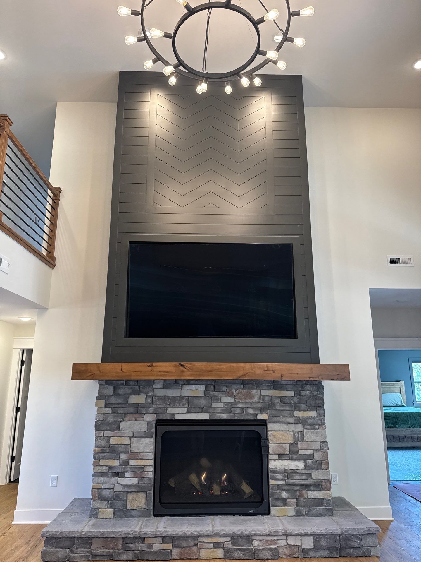 Fireplace with stone base, wooden mantel, mounted TV, gray paneling, and a chandelier.