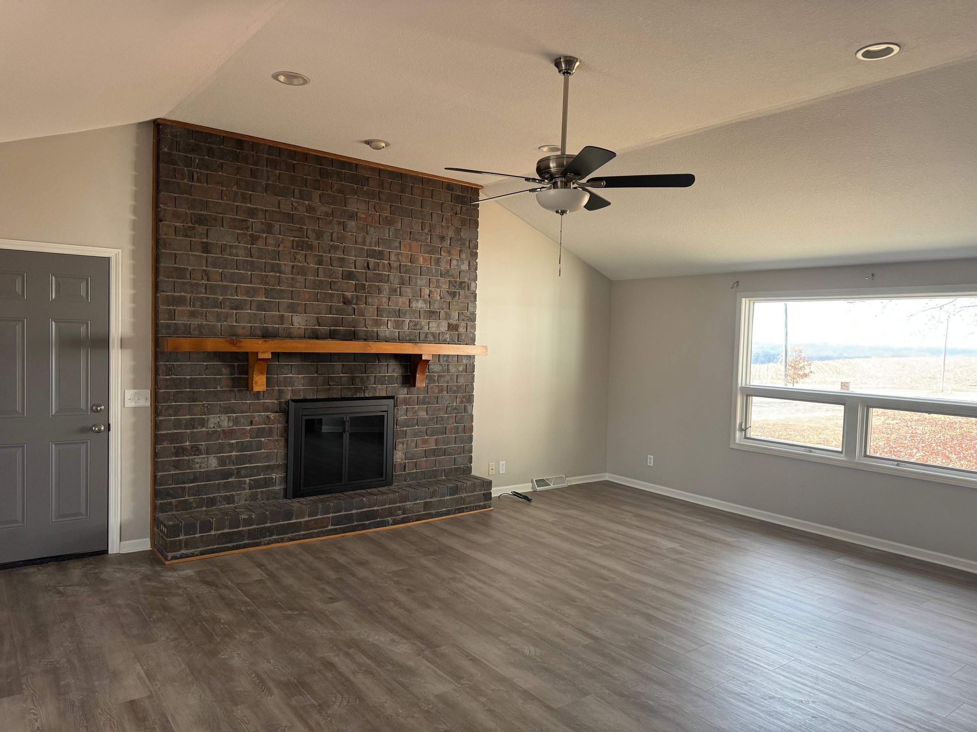 Empty living room with brick fireplace, gray walls, and wood-look flooring. Window on the right.