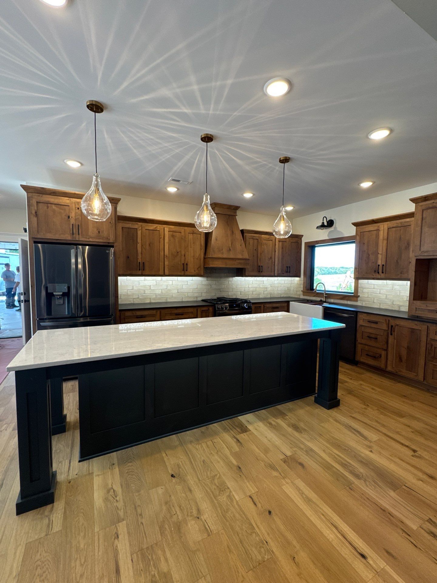 Kitchen with wooden cabinets, black island, light-colored countertops, and three hanging pendant lights.