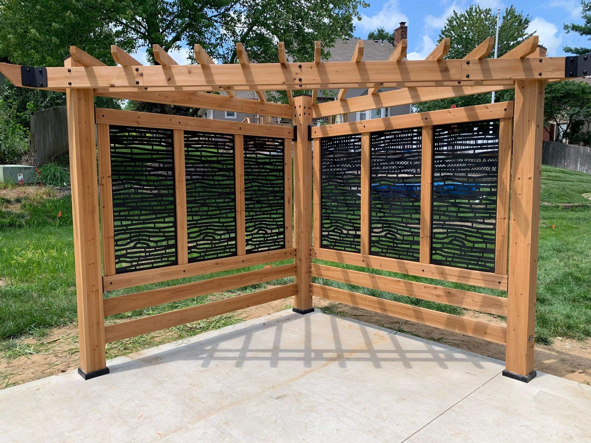 Wooden pergola with black decorative panels on a concrete patio in a grassy area.