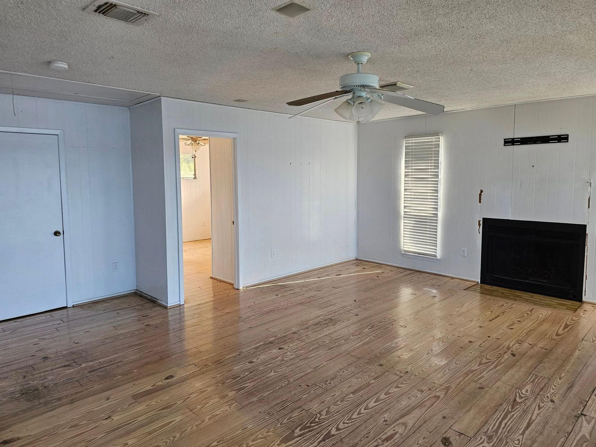 Empty living room with light wood floor, white walls, and a dark fireplace. A door and window are visible.