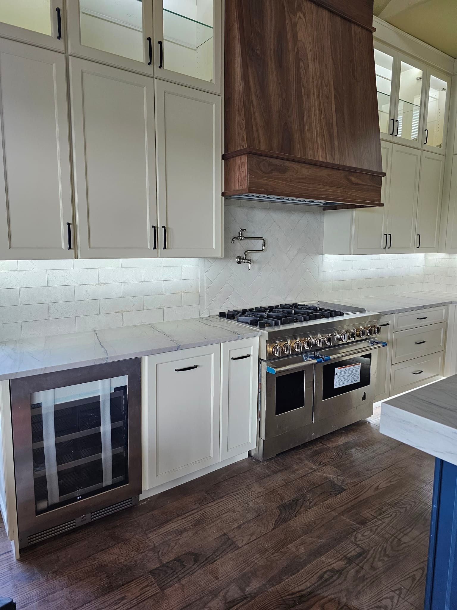A modern kitchen with white cabinets, a stainless steel range, wine cooler, and dark wood range hood. Dark wood flooring and white subway tile backsplash.