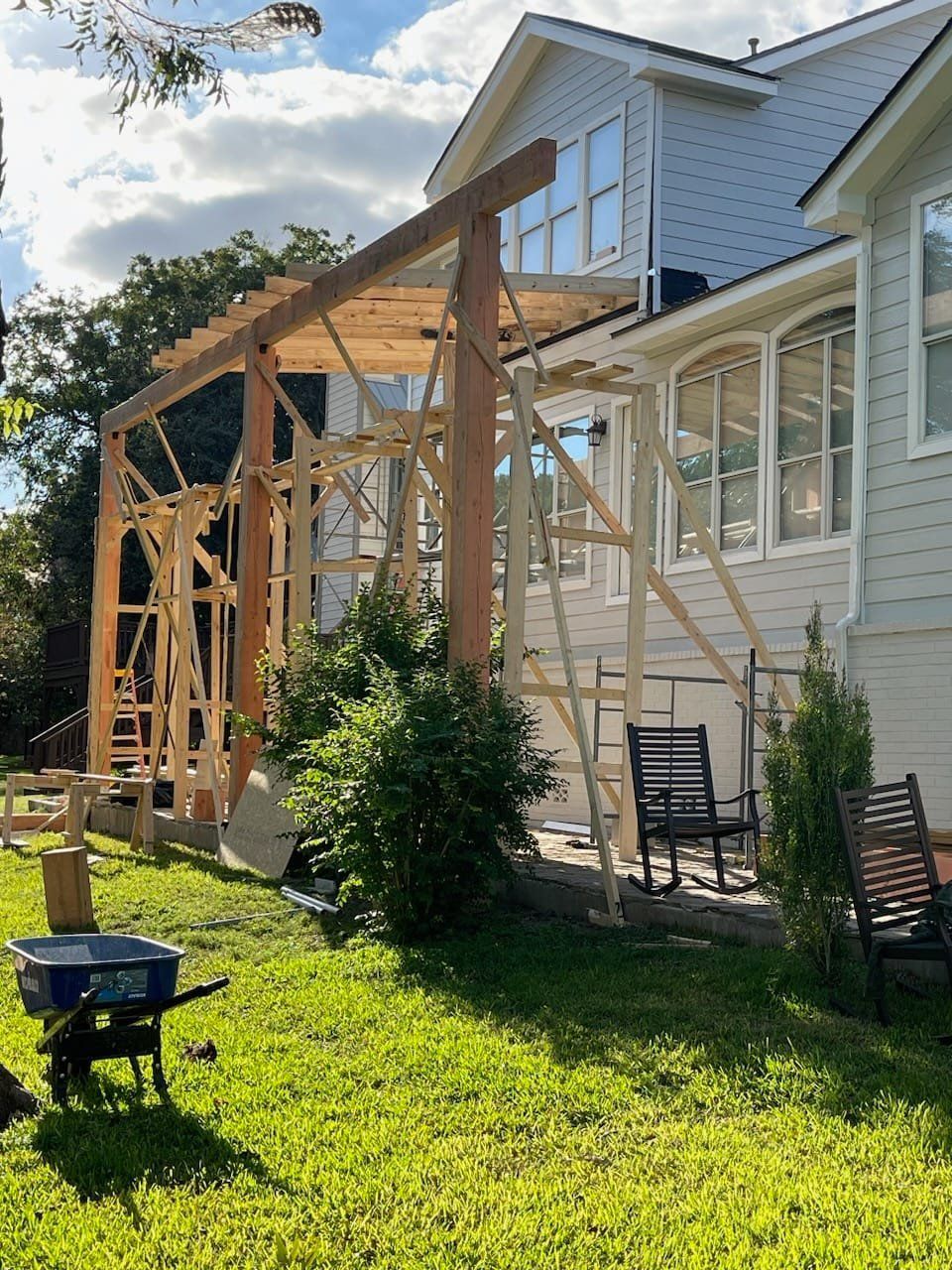 Construction of a wooden pergola attached to a two-story house with light blue siding; tools and wheelbarrow are on the green grass.